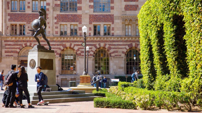 A side view of the statue, Tommy Trojan, next to a large bush with five students gather around the base chatting.