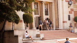Four students walking in the Physical Education Building as other students sit near the steps at the entrance answering phone calls and studying.
